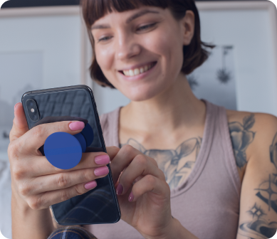 Mulher sorridente com tatuagens, usando um smartphone com suporte na parte traseira, representando uma experiência descontraída e prática ao interagir com o aplicativo indicaz.