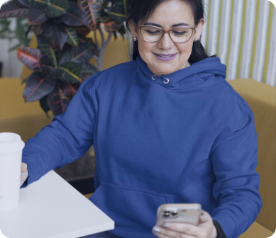 Mulher sorridente usando um moletom azul, segurando um smartphone enquanto toma um café em um ambiente aconchegante com plantas ao fundo, simbolizando praticidade e conforto ao usar o aplicativo indicaz.